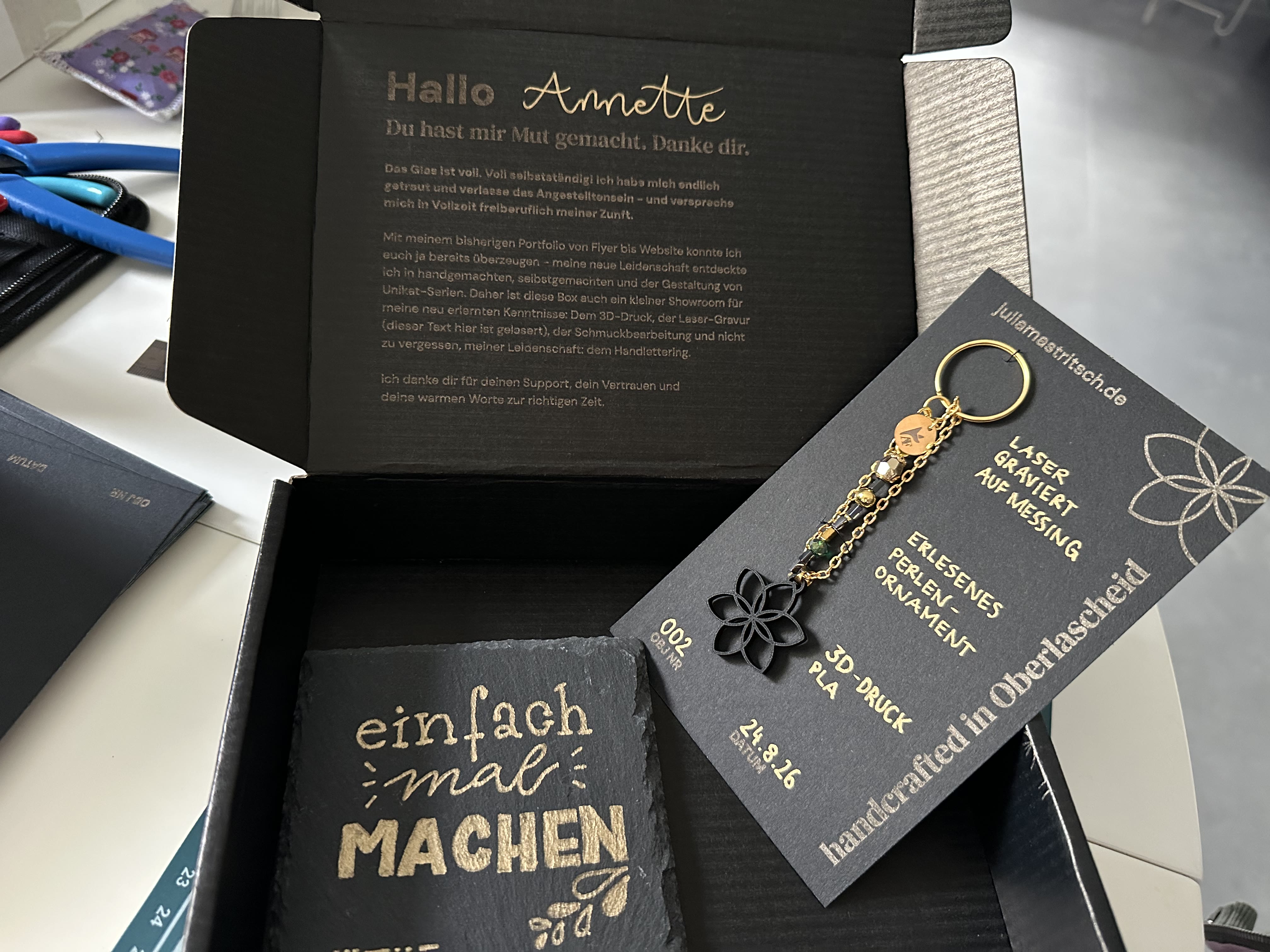 Geöffnete Box mit Grußformel Hallo Annette im Innendeckel, Schiefer-Untersetzer und Schlüsselanhänger auf Backkarte